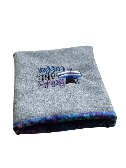 The embroidered pattern to the front of the handmade Book Pouch is 'Books and Coffee' with stacked books and a coffee cup.  The embroidered pattern is in colours of purple, blue and black.  The Book Pouch is lined with complementing blue and purple patterned fabric.  Please note the Book Pouch is open and does not have a closure.